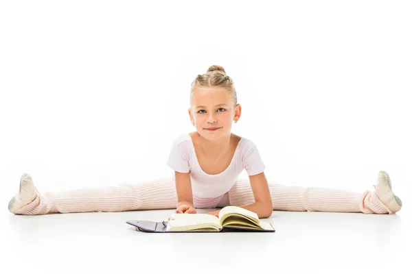 Усміхнена маленька балерина читає книгу і робить шпагат ізольовано на білому тлі — стокове фото