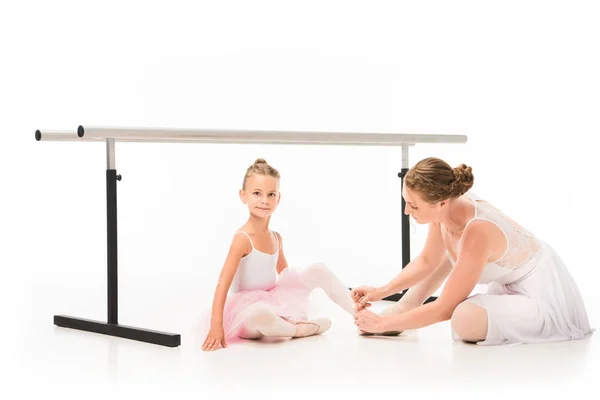 Mujer entrenador atando zapatos puntiagudos de pequeña bailarina cerca de soporte de barra de ballet aislado sobre fondo blanco - foto de stock