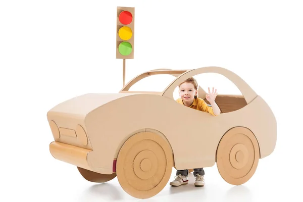 Adorable boy waving and playing with cardboard car and traffic lights on white — Stock Photo