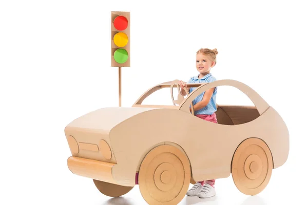 Stylish female preschooler driving cardboard car on white with traffic lights — Stock Photo