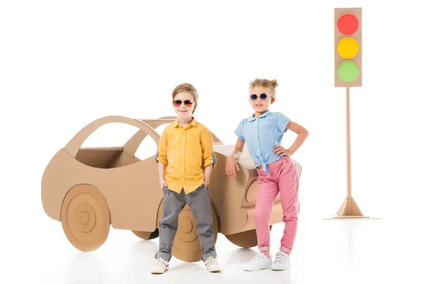 Élégants frères et sœurs adorables dans des lunettes de soleil posant ensemble près de voiture en carton et feux de circulation, sur blanc — Photo de stock