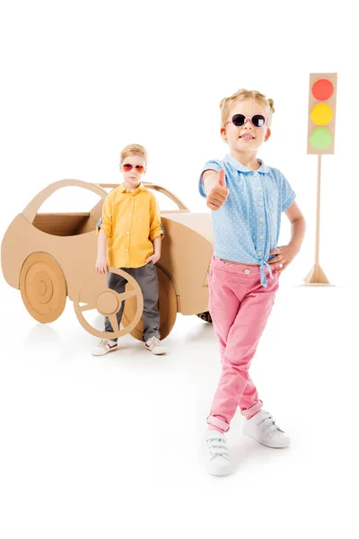 Stylish kid in sunglasses showing thumb up while boy standing near cardboard car and traffic lights, on white — Stock Photo