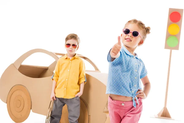 Adorable enfant élégant dans des lunettes de soleil montrant pouce levé tandis que le garçon debout près de voiture en carton et feux de signalisation, sur blanc — Photo de stock