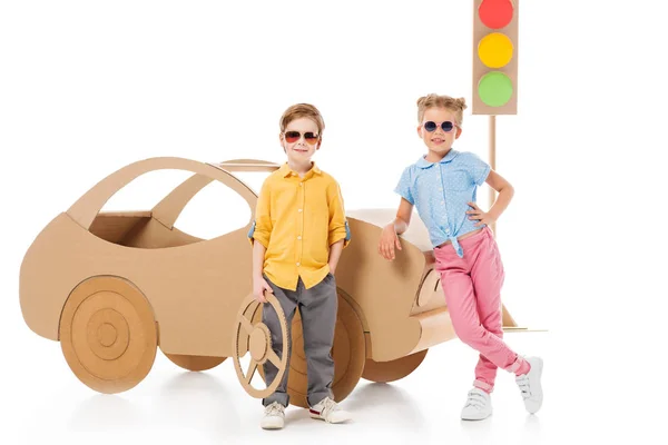 Crianças elegantes em óculos de sol posando perto de carro de papelão e semáforos, em branco — Fotografia de Stock
