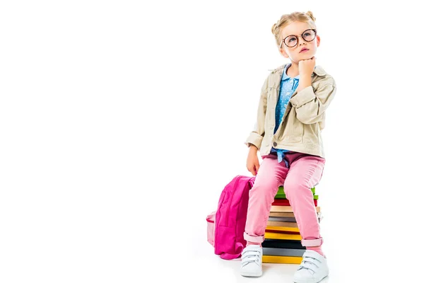 Thoughtful schoolgirl sitting on pile of books with pink backpack isolated on white — Stock Photo