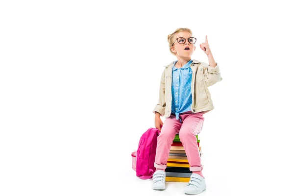 Niño escolar emocionado apuntando hacia arriba y teniendo idea mientras está sentado en un montón de libros con mochila aislada en blanco, estudiando concepto - foto de stock