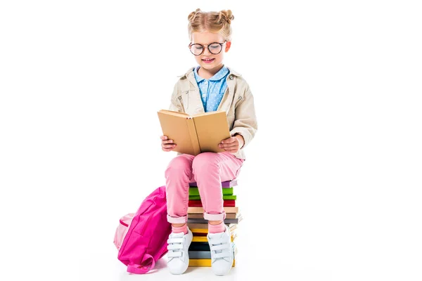 Entzückendes Schulmädchen liest Buch, während es auf einem Stapel Bücher sitzt, mit Rucksack isoliert auf weißem, pädagogischem Konzept — Stockfoto