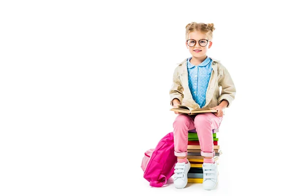 Adorable pupil reading while sitting on pile of books with pink backpack isolated on white — Stock Photo