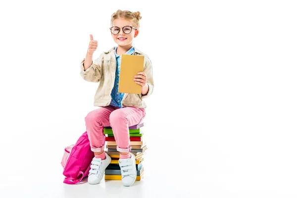 Adorabile allievo sorridente mostrando pollice in su mentre seduto su mucchio di libri isolati su bianco, concetto di conoscenza — Foto stock