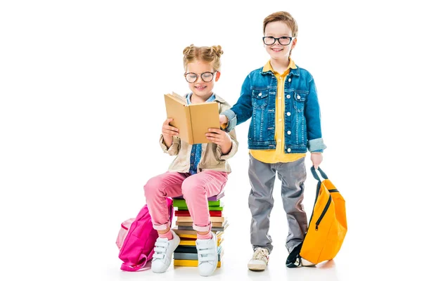 Schoolchildren in eyeglasses studying and sitting on books with backpacks, isolated on white — Stock Photo