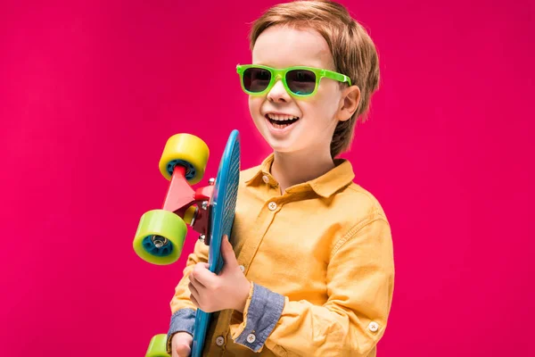Стильний усміхнений хлопчик в сонцезахисних окулярах позує зі скейтбордом ізольовано на червоному — стокове фото