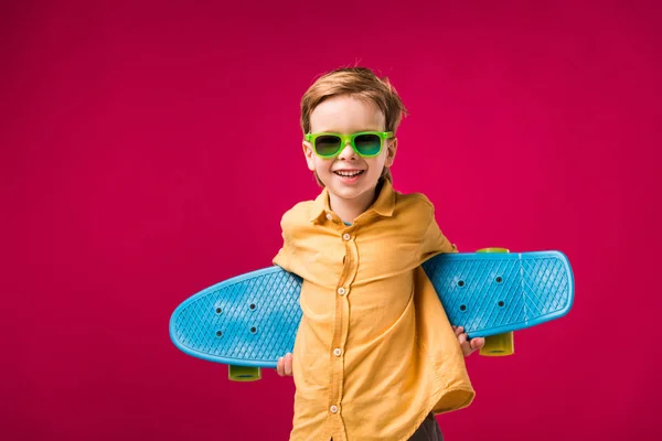 Stylish cheerful boy in sunglasses posing with penny board isolated on red — Stock Photo