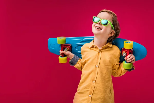 Adorable sonriente pequeño patinador en gafas de sol posando con penny board aislado en rojo - foto de stock