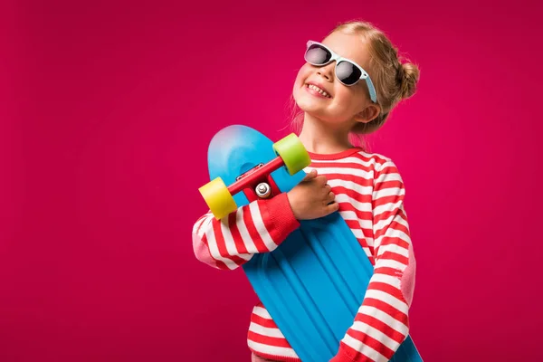 Niño con estilo feliz en gafas de sol posando con monopatín aislado en rojo - foto de stock
