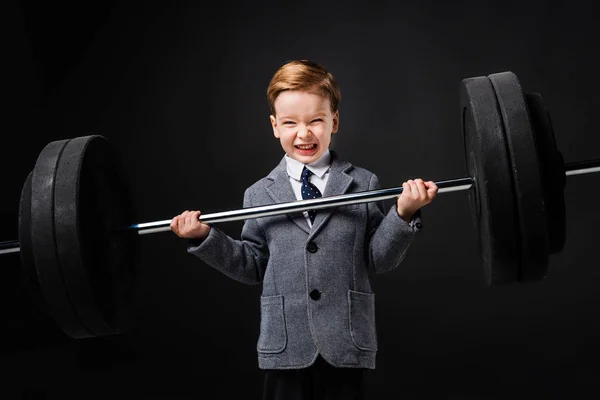 Strong little boy in suit lifting barbell isolated on grey — Stock Photo