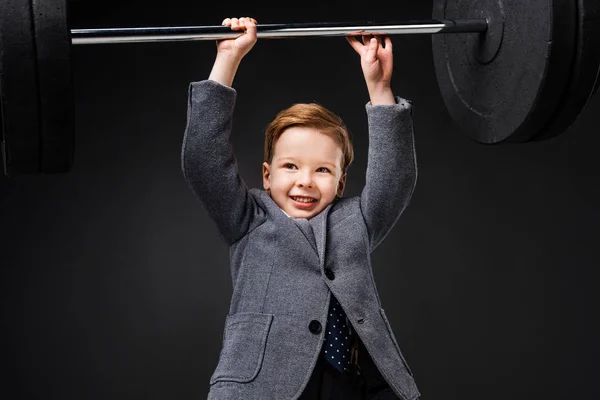 Сильный улыбающийся мальчик в костюме поднимающий штангу изолированный на сером — стоковое фото