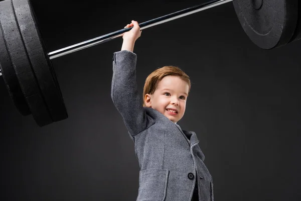 Сильный веселый мальчик в костюме поднимая штангу изолированы на серый — стоковое фото