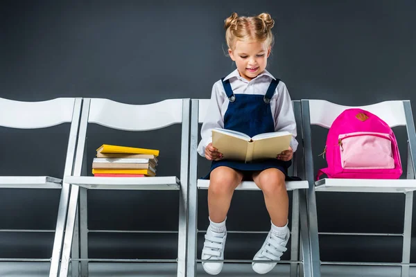 Entzückendes Schulkind liest und sitzt mit Büchern und Rucksack auf Stühlen — Stockfoto