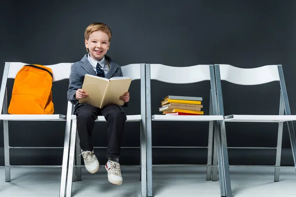 Lächelnder Schuljunge liest und sitzt mit Büchern und Rucksack auf Stühlen — Stockfoto