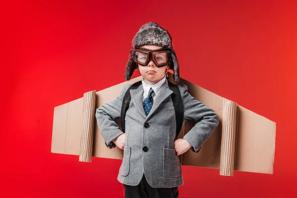 Niño molesto en traje con alas de avión y gafas protectoras aisladas en rojo - foto de stock