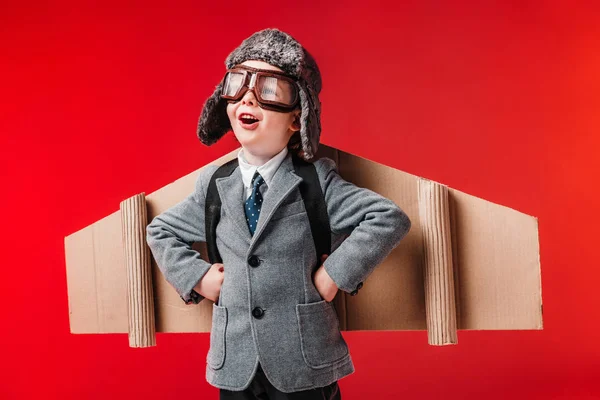 Niño excitado en traje con alas de avión de papel y gafas aisladas en rojo - foto de stock