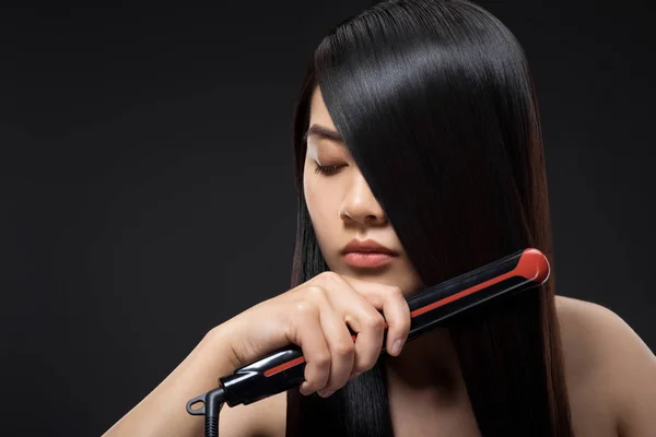 Portrait of young asian woman straightening hair with hair straightener isolated on black — Stock Photo