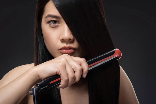 Portrait of young asian woman straightening hair with hair straightener isolated on black — Stock Photo