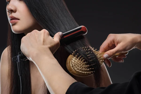 Vue partielle du coiffeur lissant womans cheveux avec lisseur et brosse isolée sur noir — Photo de stock