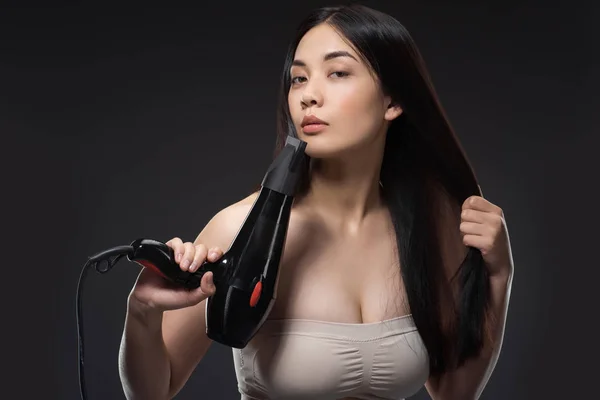 Portrait of attractive asian woman with hair dryer isolated on black — Stock Photo