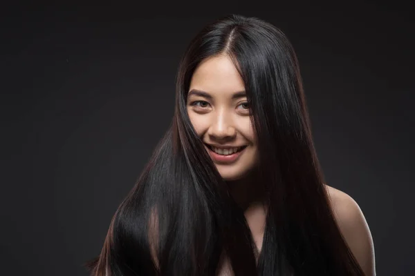 Retrato de joven sonriente mujer asiática con hermoso y saludable cabello oscuro mirando a la cámara aislada en negro - foto de stock
