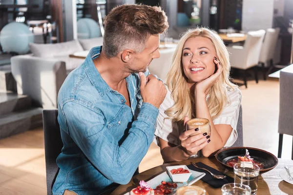 Hermosa novia rubia sosteniendo taza de café y mirando novio en la mesa en la cafetería - foto de stock