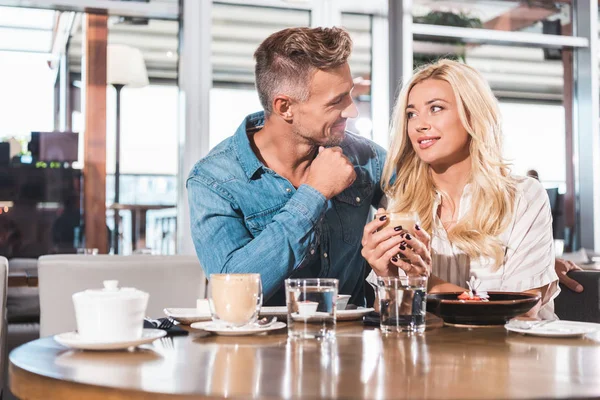 Cheerful girlfriend holding cup of coffee and looking at handsome boyfriend at table in cafe — Stock Photo