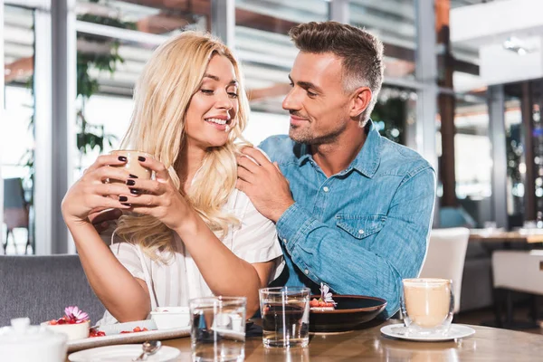 Boyfriend hugging smiling girlfriend and she holding cup of coffee at table in cafe — Stock Photo