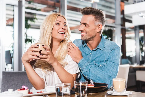 Boyfriend hugging laughing girlfriend and she holding cup of coffee at table in cafe — Stock Photo