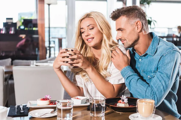 Freund umarmt fröhliche Freundin und sie hält Tasse Kaffee am Tisch im Café — Stockfoto