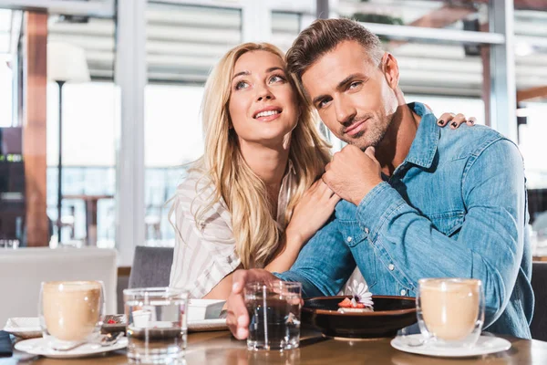 Подружка обнимает бойфренда и он смотрит в камеру за столиком в кафе — стоковое фото