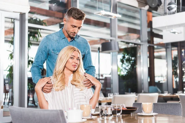 Handsome boyfriend standing near table and hugging attractive girlfriend in cafe — Stock Photo