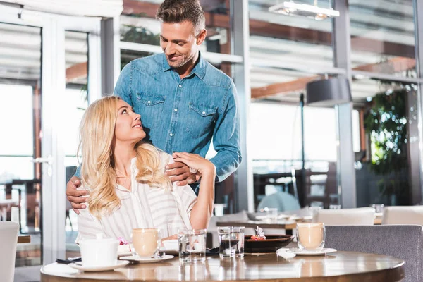 Novio guapo de pie cerca de la mesa, abrazando a la novia y se miran en la cafetería - foto de stock