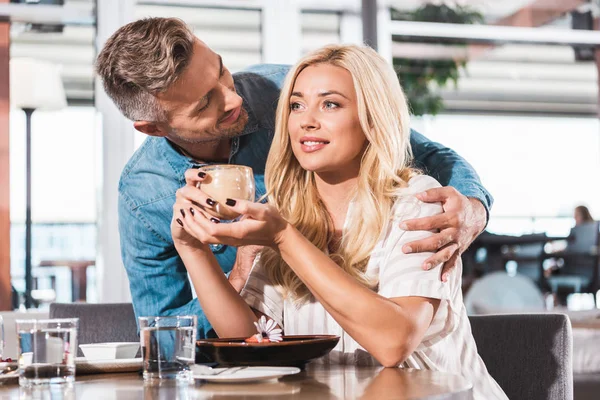 Bonito namorado abraçando namorada perto de mesa no café, ela segurando copo de café — Fotografia de Stock