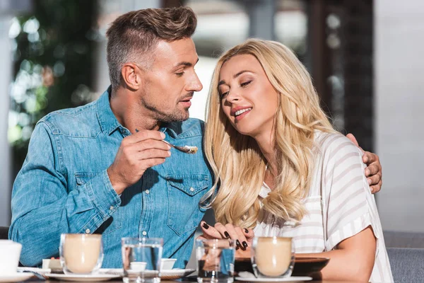 Guapo novio alimentación sonriente novia con sabroso postre durante la fecha en la cafetería - foto de stock