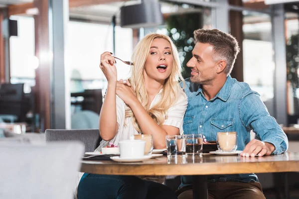 Happy girlfriend grimacing and feeding boyfriend with tasty dessert at table in cafe — Stock Photo