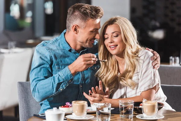 Happy boyfriend feeding attractive girlfriend with sweet dessert at table in cafe — Stock Photo