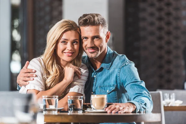 Happy tender heterosexual couple hugging at table in cafe and looking at camera — Stock Photo