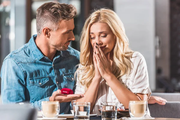 Handsome boyfriend making proposal to surprised girlfriend and holding ring box in cafe — Stock Photo