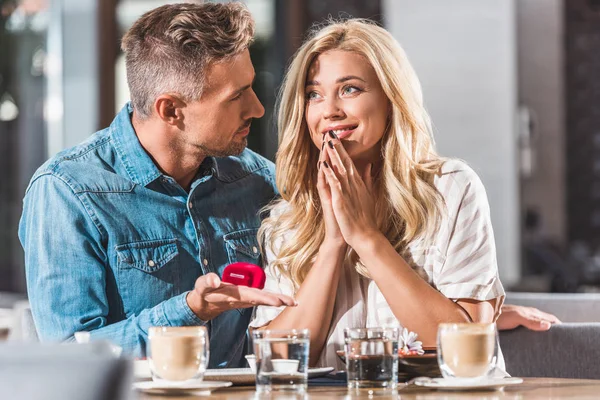 Handsome boyfriend proposing happy attractive girlfriend and holding ring box in cafe — Stock Photo