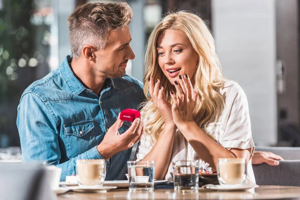 Handsome boyfriend proposing shocked girlfriend and showing red ring box in restaurant — Stock Photo