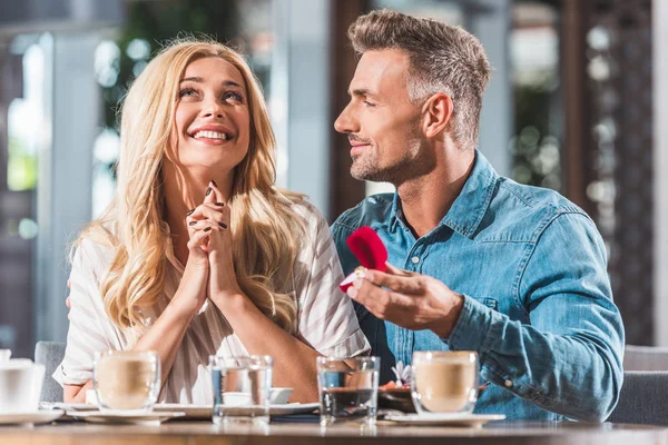 Cheerful handsome boyfriend proposing surprised girlfriend and holding ring box in cafe — Stock Photo