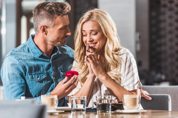 Handsome boyfriend proposing surprised attractive girlfriend and holding ring box in cafe — Stock Photo