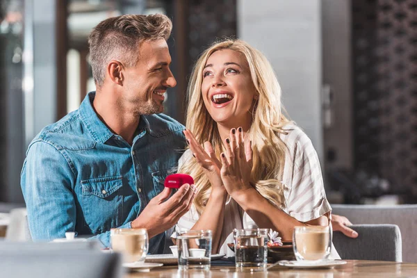 Beau petit ami proposant petite amie geste choqué et tenant boîte à bague dans le café — Photo de stock