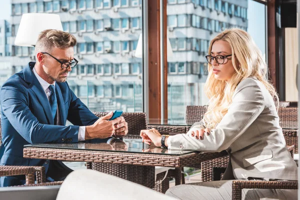 Mujer de negocios comprobar el tiempo y hombre de negocios utilizando el teléfono inteligente en la mesa en la cafetería - foto de stock